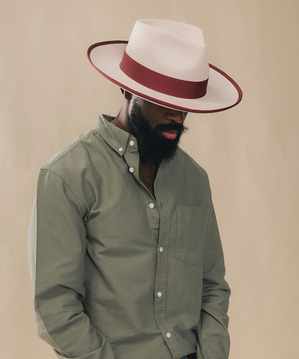 A man wearing a white fedora hat with a dark red band and a muted sage green button-down shirt, posed against a solid beige background #color_maroon