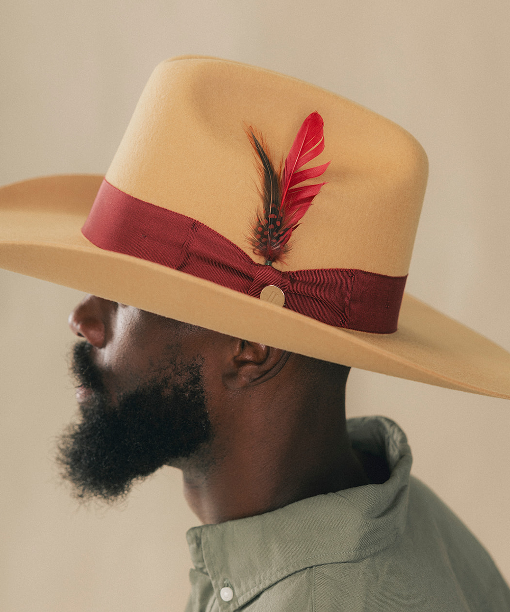 A close-up side profile of a man wearing a wide-brim fedora hat in light tan with a burgundy ribbon band and a red feather accent, against a plain beige background #color_mustard/maroon