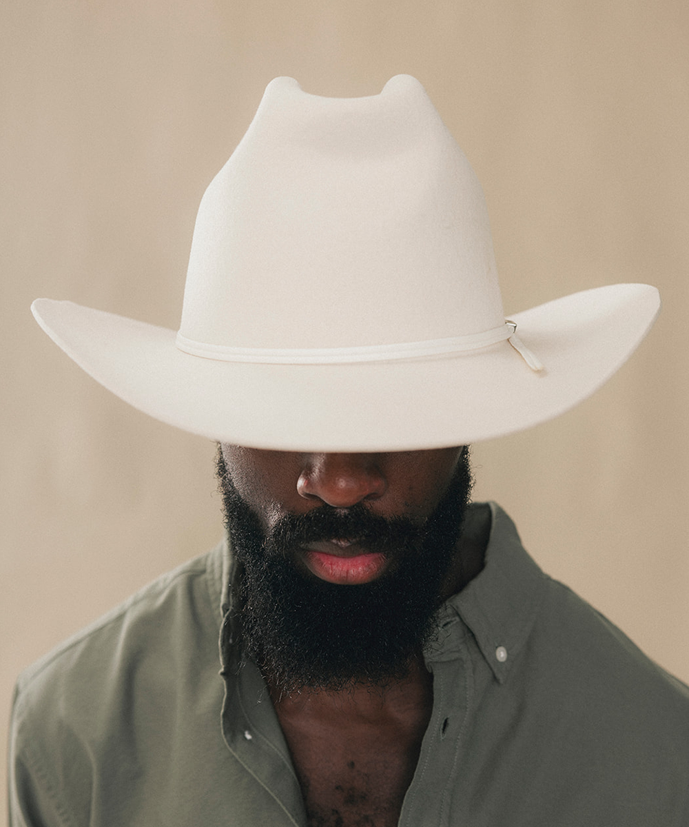 A man with a dark beard wearing a white cowboy hat and an olive green button-down shirt, shown from the chest up against a plain beige background #color_bone