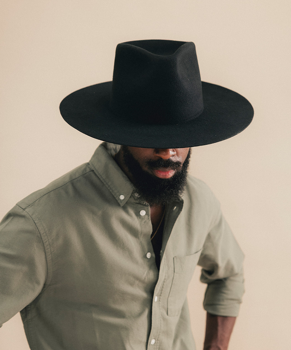 A man wearing a wide-brimmed black cowboy hat and a muted sage green button-down shirt, photographed against a plain beige studio background #color_black
