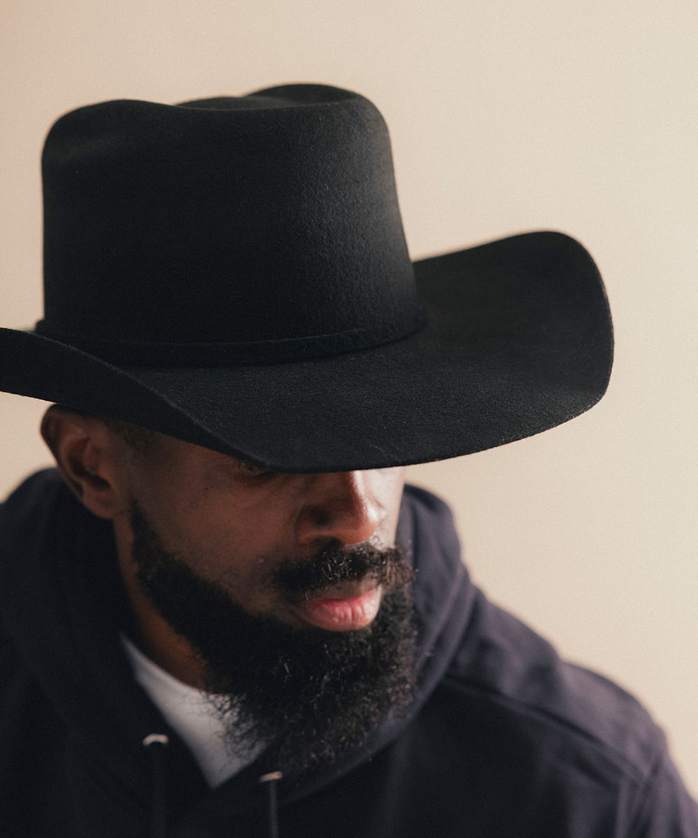 A close-up portrait of a man wearing a black felt cowboy hat and a dark hoodie against a plain beige background #color_black