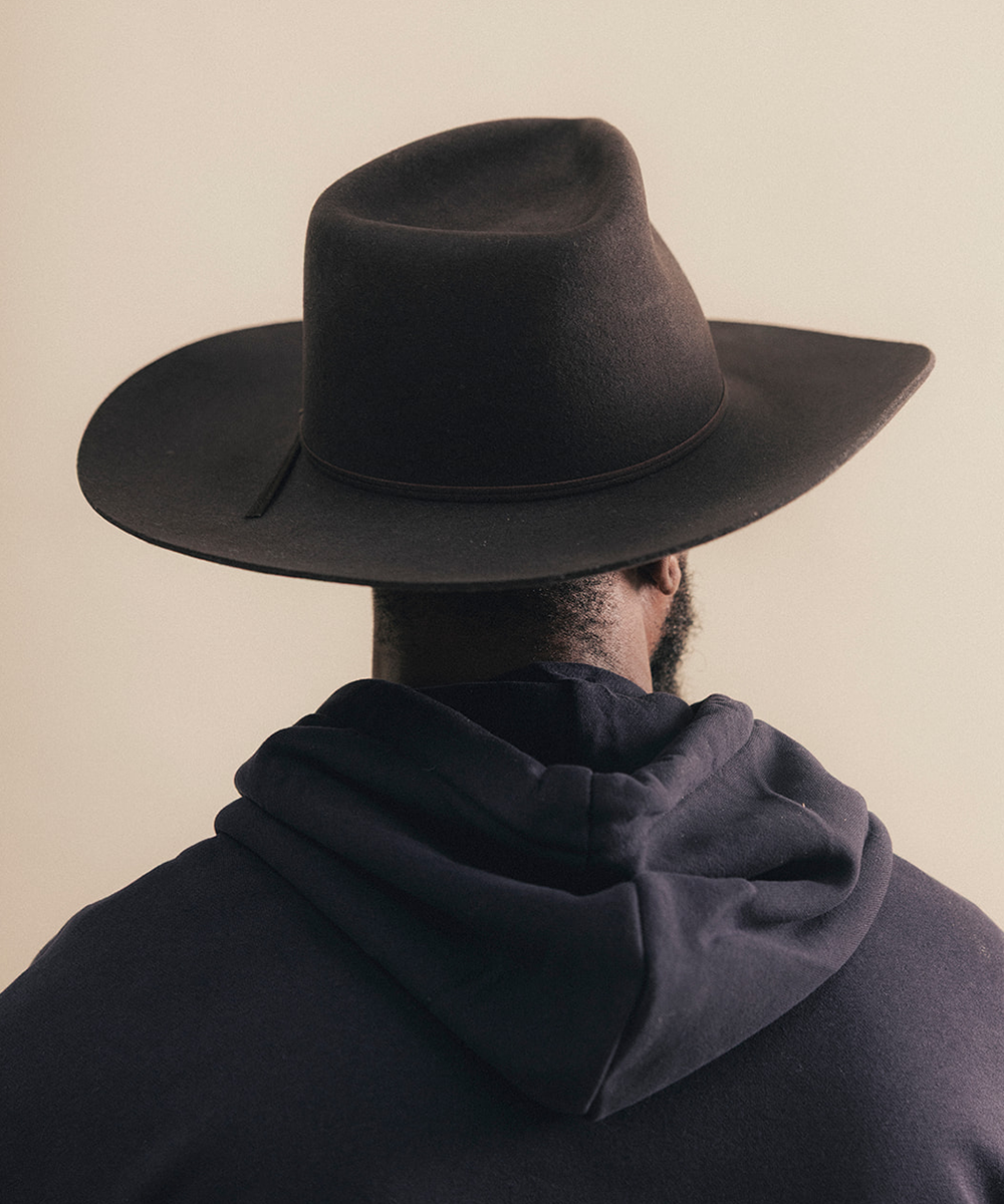 A man viewed from behind wearing a wide-brimmed black felt cowboy hat and a dark navy hoodie, set against a plain beige background #color_hickory
