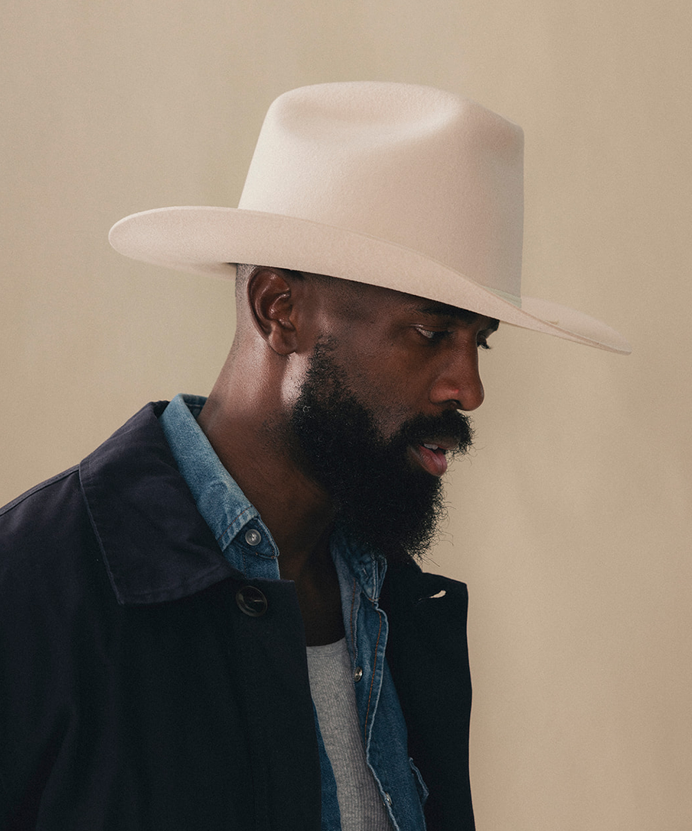A man with a dark beard wearing a light cream-colored cowboy hat, a dark jacket, and a denim shirt, shown in profile against a solid beige background #color_buckskin