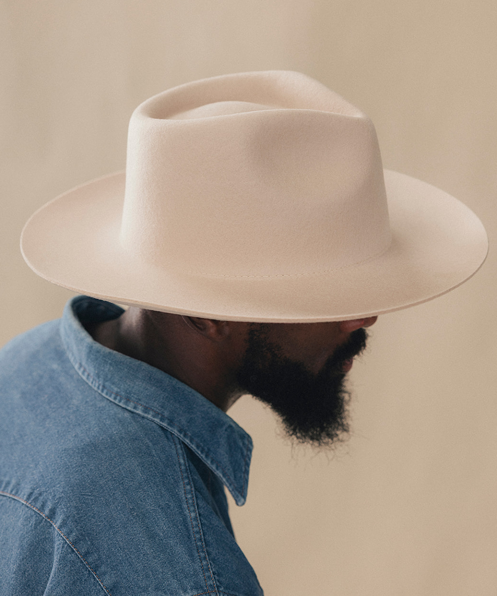 A man in profile wearing a light beige wide-brim fedora hat and a blue denim shirt, set against a solid warm beige background #color_buckskin