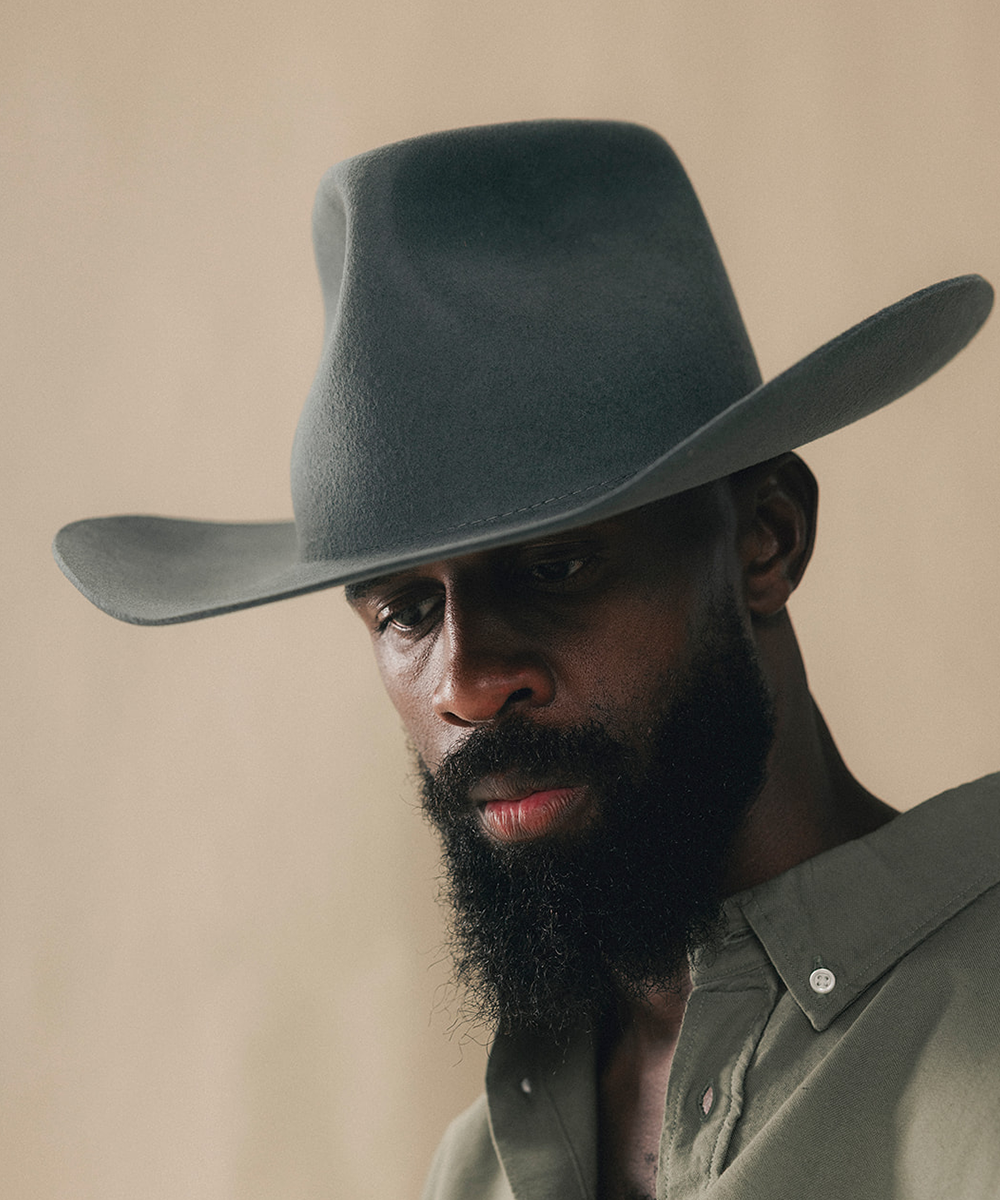 A close-up portrait of a man wearing a dark gray felt cowboy hat and a muted olive green button-up shirt against a warm beige background #color_dark grey