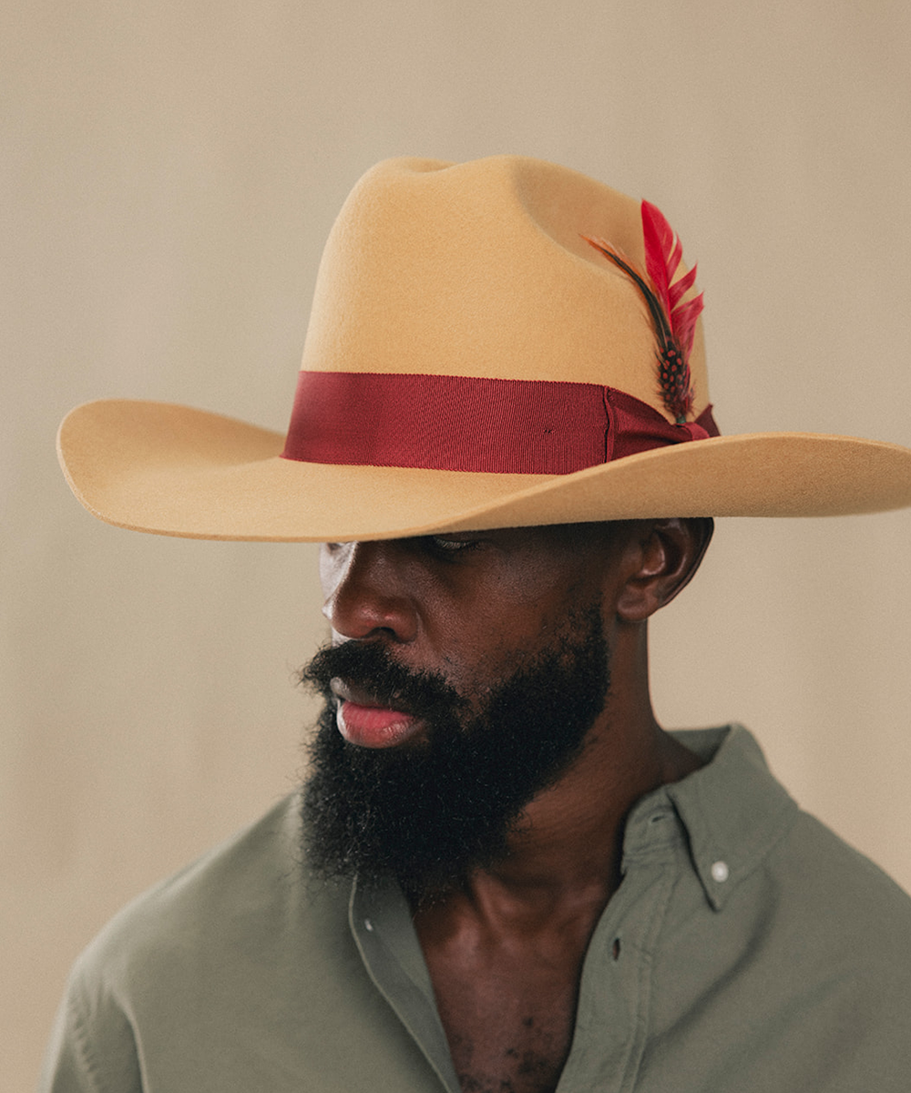 A man wearing a wide-brimmed tan cowboy hat with a dark red band and a red feather accent, paired with an olive green shirt, set against a plain beige background #color_mustard/maroon