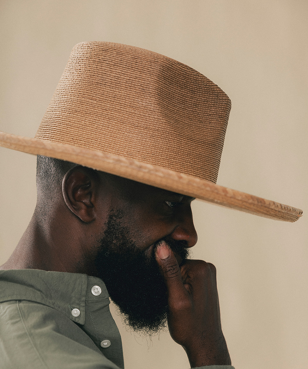 A man in profile wearing a wide-brimmed woven straw hat and an olive green shirt, set against a plain beige background #color_oak