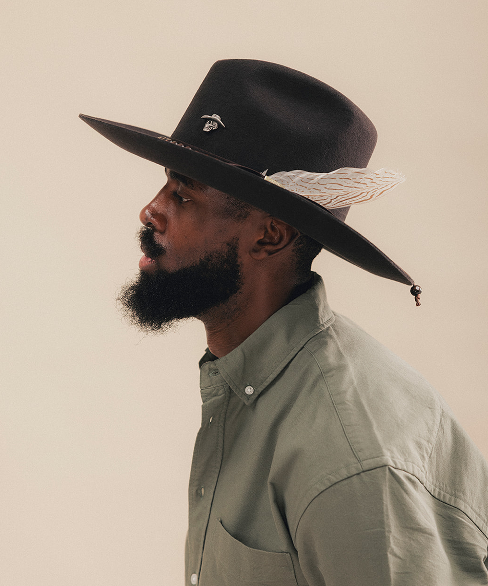 A man in profile wearing a dark brown wide-brim cowboy hat with a white feather accent and a sage green button-down shirt, set against a solid beige background #color_hickory