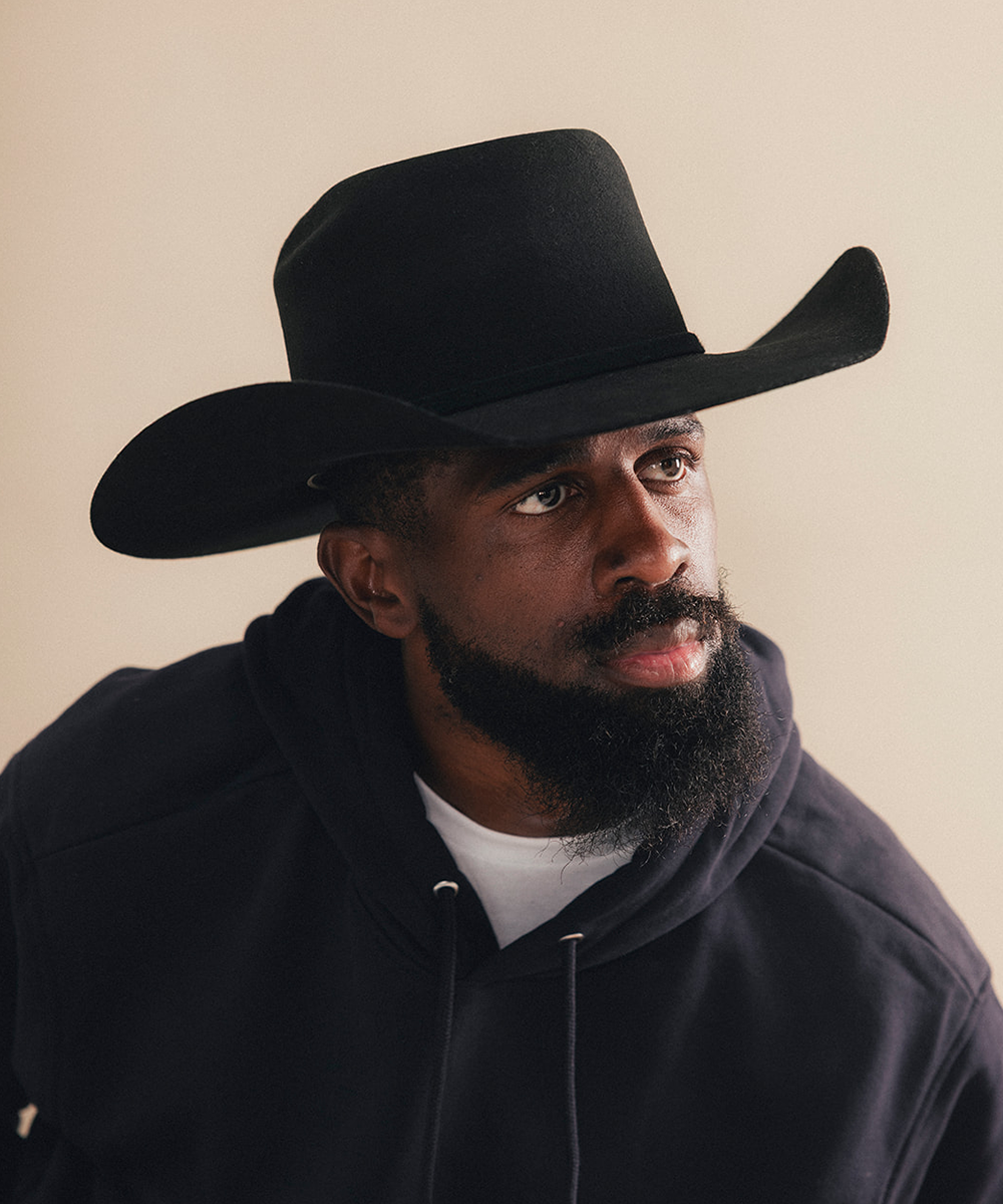 A man with a beard wearing a black cowboy hat and a black hoodie over a white t-shirt, photographed against a solid beige background #color_black