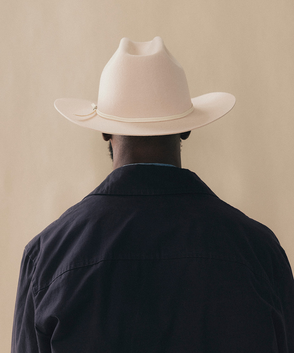 A man viewed from behind wearing a cream-colored cowboy hat with a matching band and a dark navy jacket, set against a solid beige background #color_buckskin