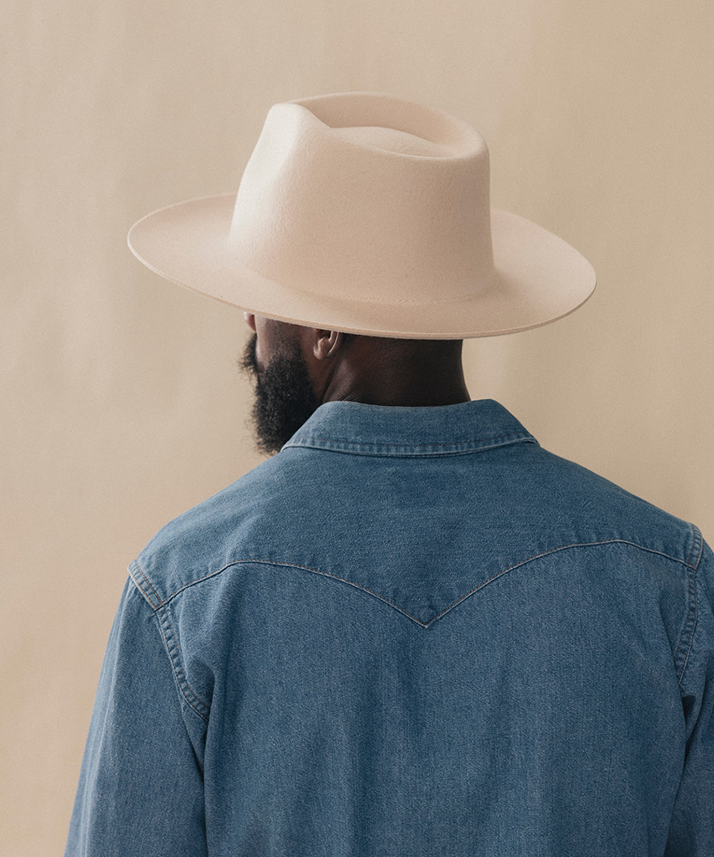 A man viewed from behind wearing a light beige wide-brim fedora hat and a blue denim shirt against a solid beige background #color_buckskin