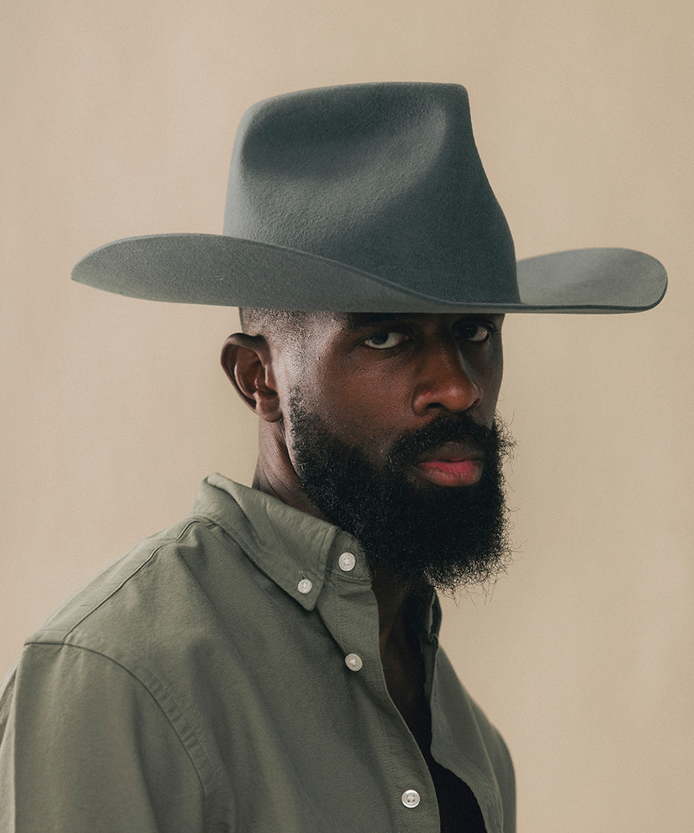 A man with a dark beard wearing a wide-brimmed dark gray cowboy hat and an olive green button-down shirt, photographed against a solid beige background #color_dark grey