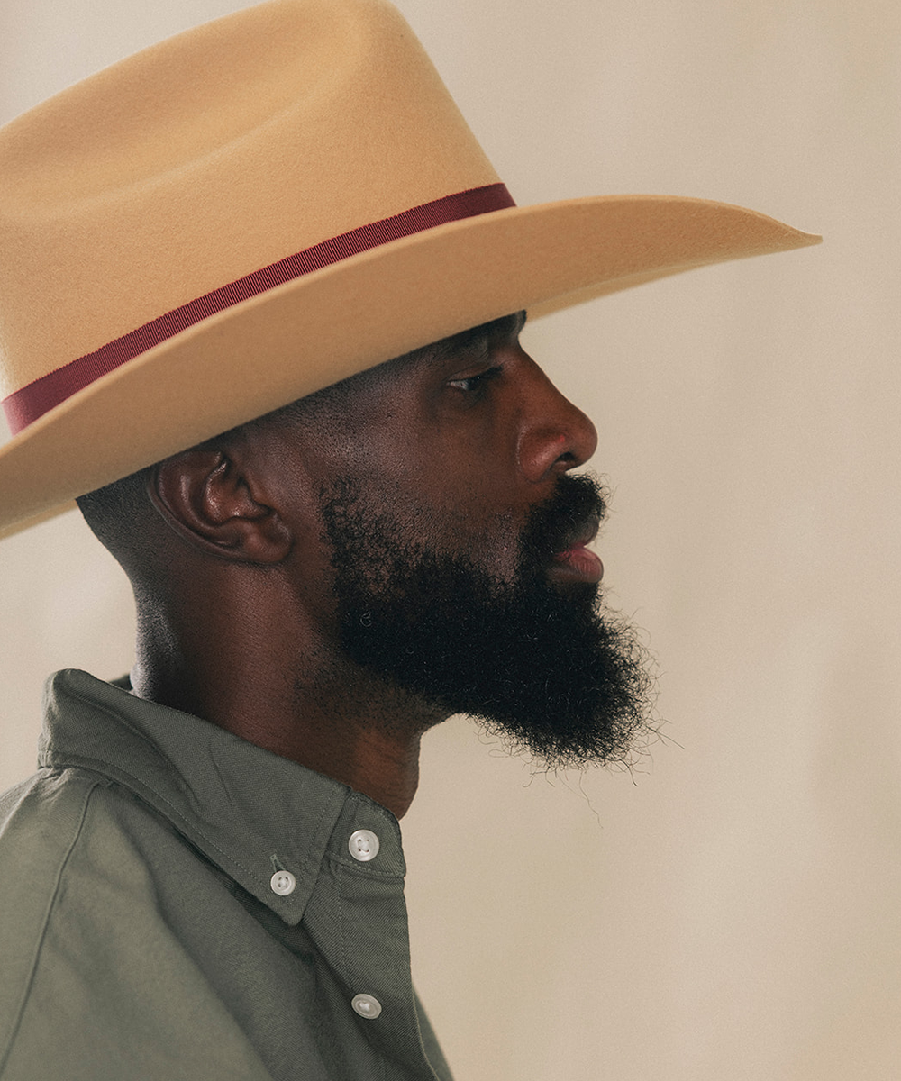 A man in profile wearing a light tan cowboy hat with a dark burgundy band and a muted green button-down shirt, set against a plain beige background #color_mustard/maroon