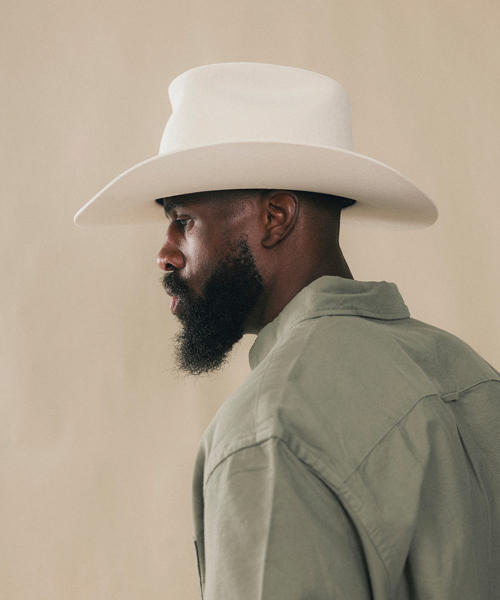 A man in profile wearing a white cowboy hat and an olive green shirt, photographed against a plain beige background #color_bone