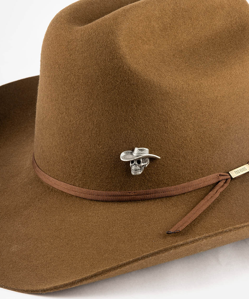 A close-up of a brown felt cowboy hat featuring a brown ribbon band and a silver metal skull pin with a small cowboy hat detail #color_anti silver