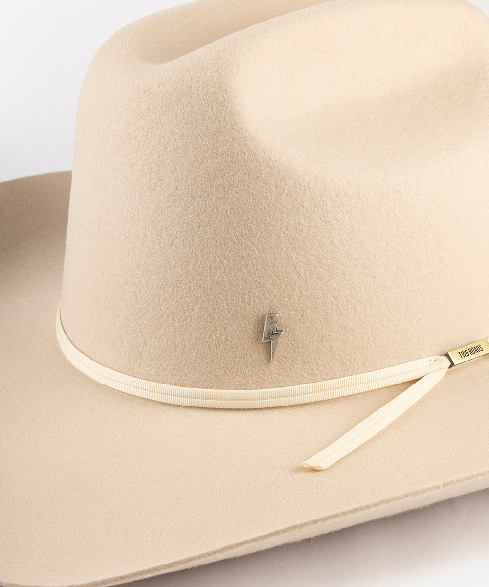 A close-up of a beige felt cowboy hat with a cream-colored band, a small gold lightning bolt pin, and a gold brand tag #color_anti silver