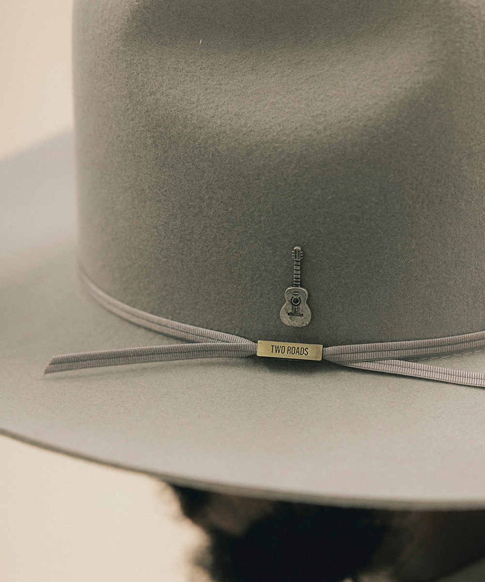 Close-up of a light gray felt hat with a thin gray ribbon band and a small guitar-shaped charm featuring a gold tag that reads 'TWO ROADS'. #color_anti silver