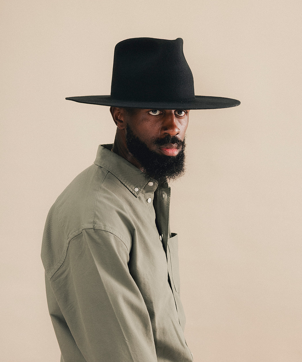 A man wearing a wide-brim black fedora hat and a light sage green button-down shirt, photographed against a solid beige background #color_black