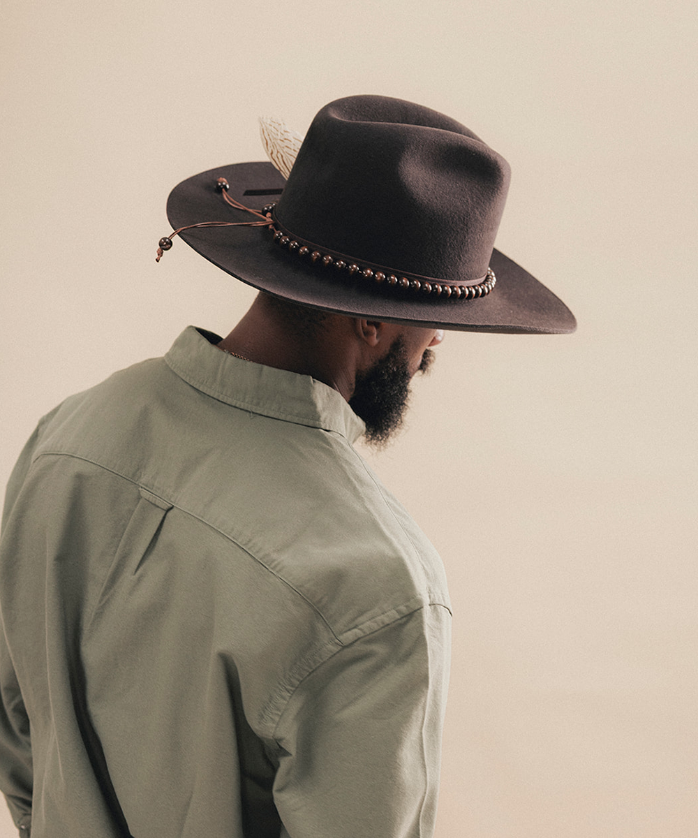 A man shown from the back wearing a dark brown wide-brim fedora hat with a decorative band and feather, paired with an olive green shirt against a plain beige background #color_hickory