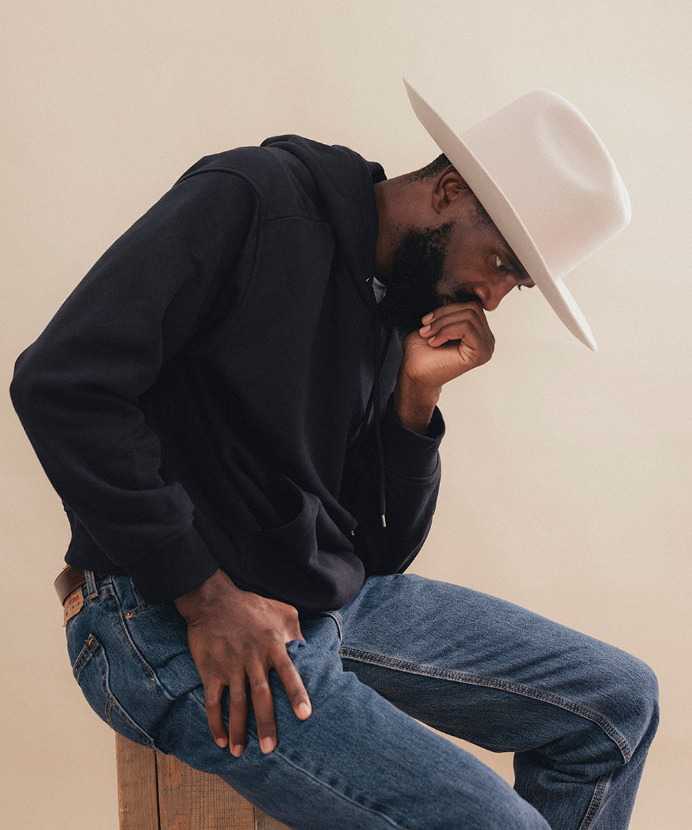 A man wearing a black hoodie, blue jeans, and a white wide-brim hat sits on a wooden stool against a plain beige background #color_silverbelly