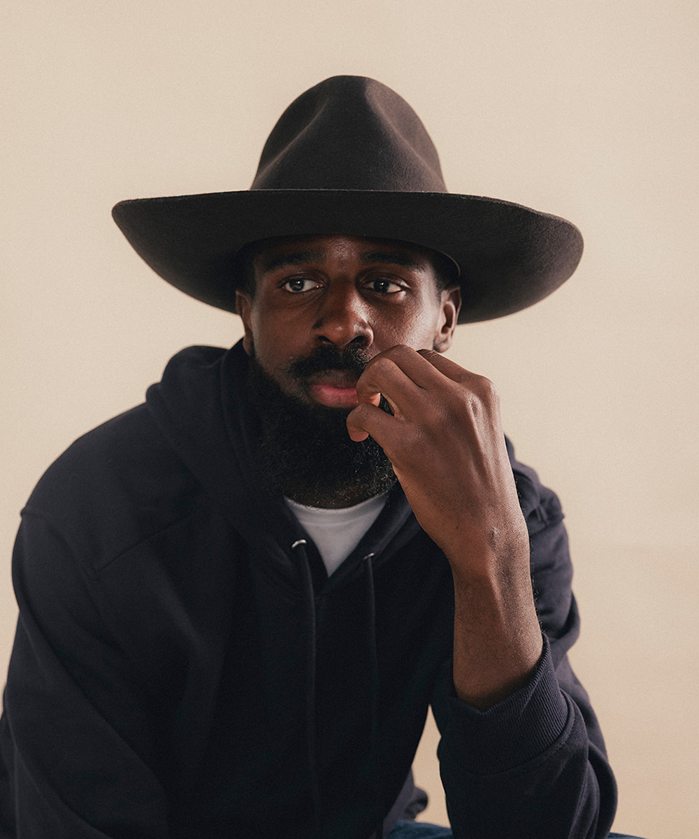 A man wearing a wide-brimmed dark brown hat and a black hoodie over a white t-shirt, posing against a solid beige background #color_hickory