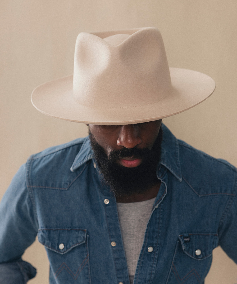 A man wearing a wide-brimmed beige fedora hat and a blue denim shirt over a gray t-shirt, photographed against a warm beige background #color_buckskin