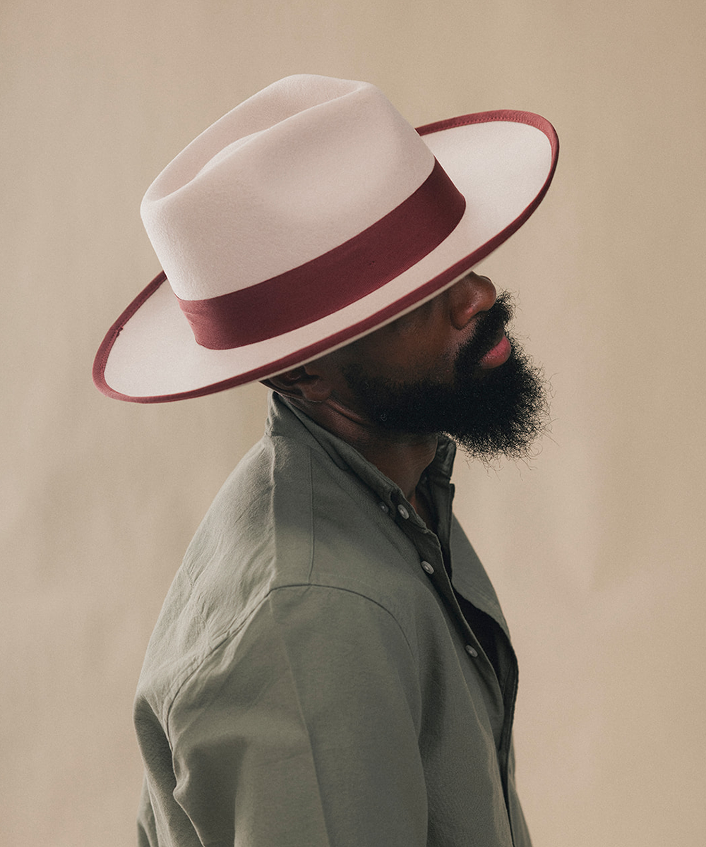 A man in profile wearing a white fedora hat with a burgundy band and matching trim, paired with a muted green shirt, against a plain beige background #color_maroon