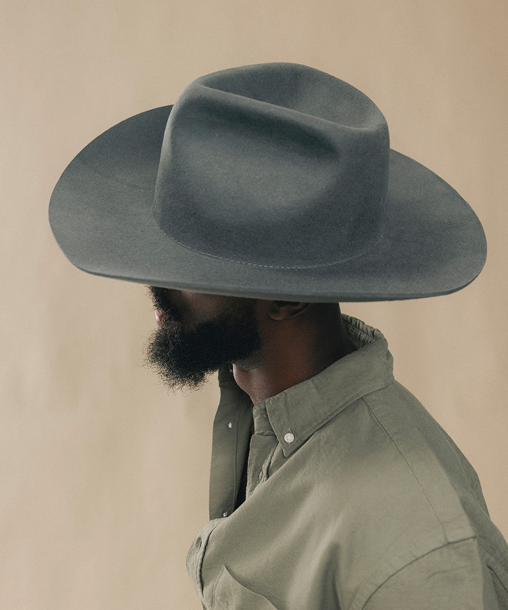 A man in profile wearing a wide-brimmed dark gray felt cowboy hat and a muted sage green button-down shirt, set against a solid beige background #color_dark grey