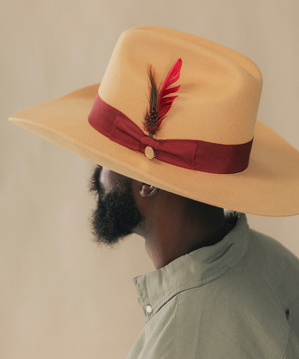 A man in profile wearing a wide-brimmed tan hat with a dark red ribbon band and a red feather accent, against a plain beige background #color_mustard/maroon