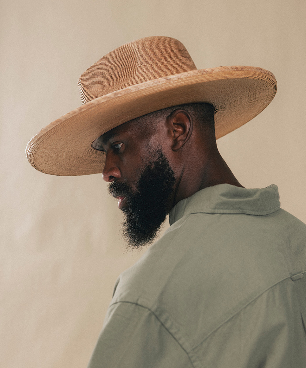 A man in profile wearing a wide-brimmed natural straw hat and a light sage green shirt, photographed against a plain beige background #color_oak