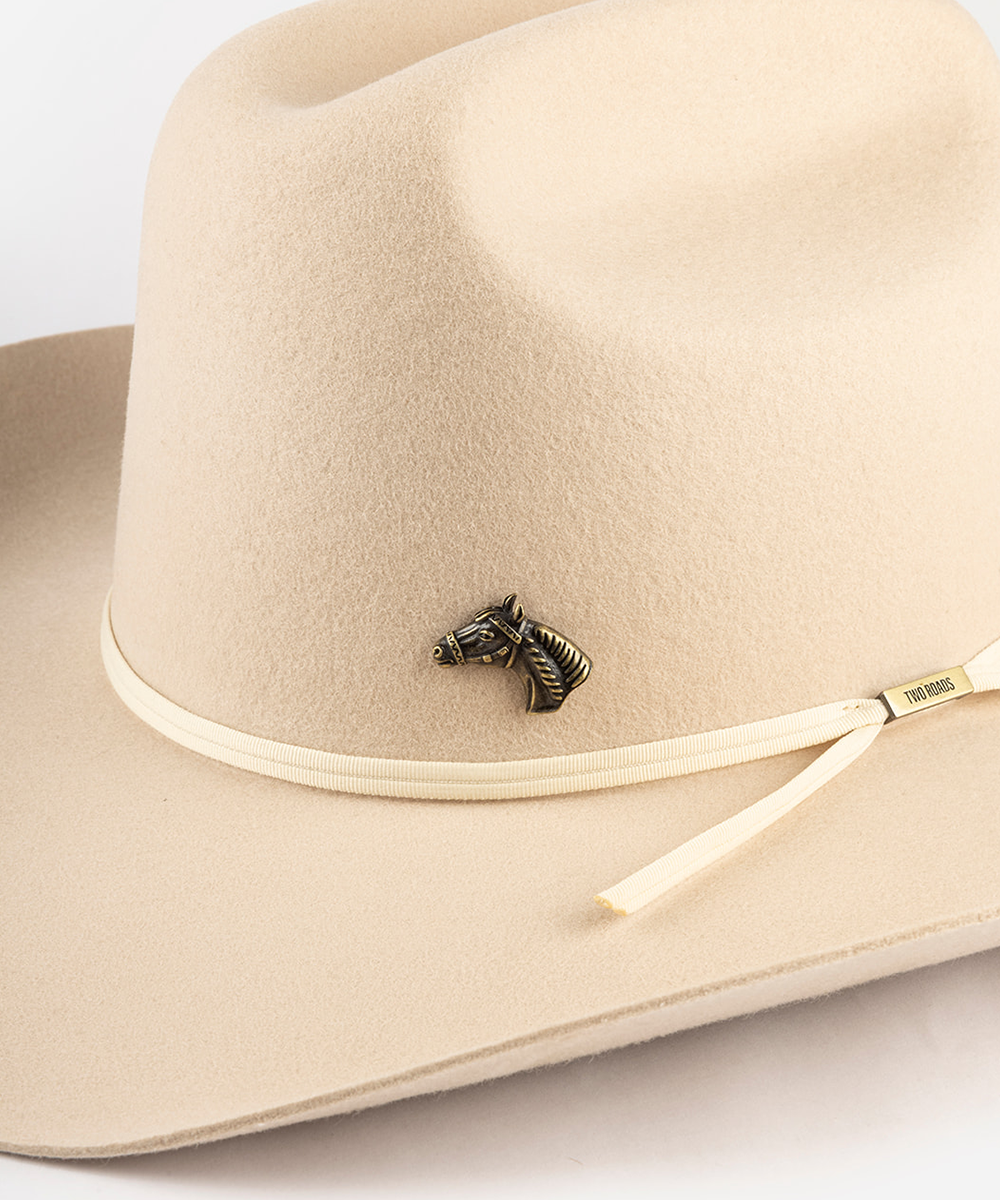 A close-up of a beige felt cowboy hat featuring a cream-colored band with a small black horse-head pin and a gold-toned tip, set against a clean white background #color_anti gold