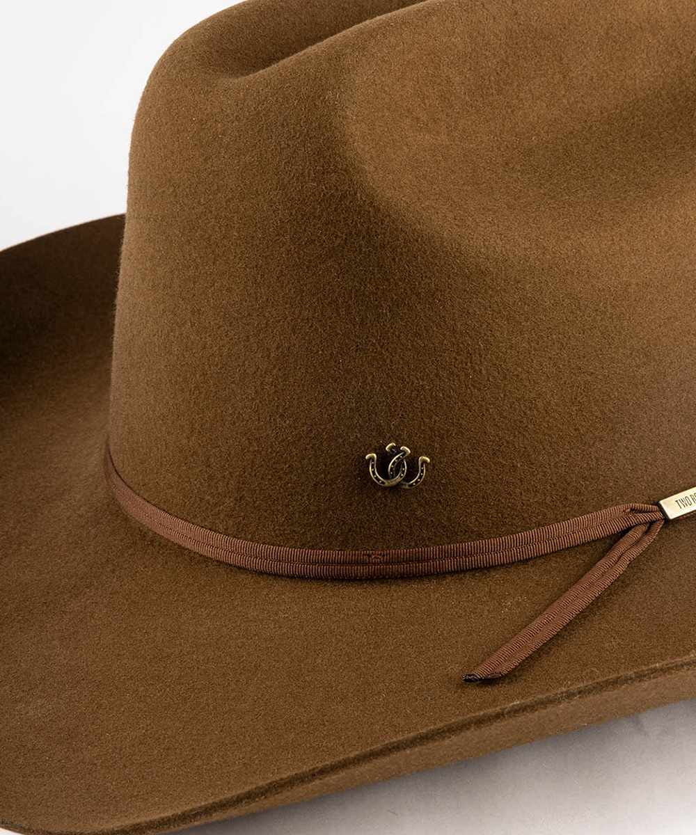 A close-up of a brown felt cowboy hat featuring a matching brown ribbon band with a small gold-toned decorative emblem and a small white brand tag #color_anti gold