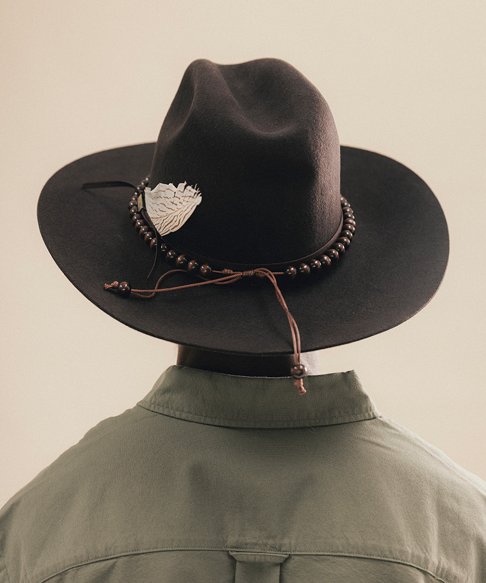 A black felt cowboy hat with a brown beaded band, silver-toned decorative emblem, and brown cord tie, shown on a mannequin wearing an olive green shirt against a warm beige background #color_hickory