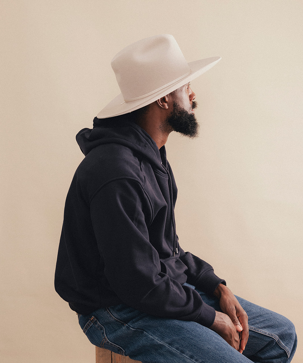 A man in profile wearing a black hoodie, blue jeans, and a cream-colored wide-brim hat, seated against a plain beige background #color_silverbelly