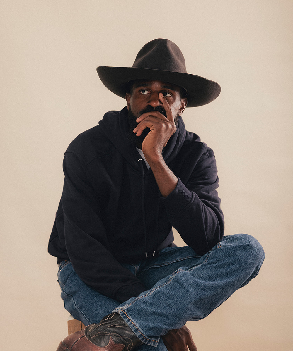 A man wearing a black wide-brim hat, black hoodie, blue jeans, and brown boots, sitting in a crouched pose against a solid beige background #color_hickory