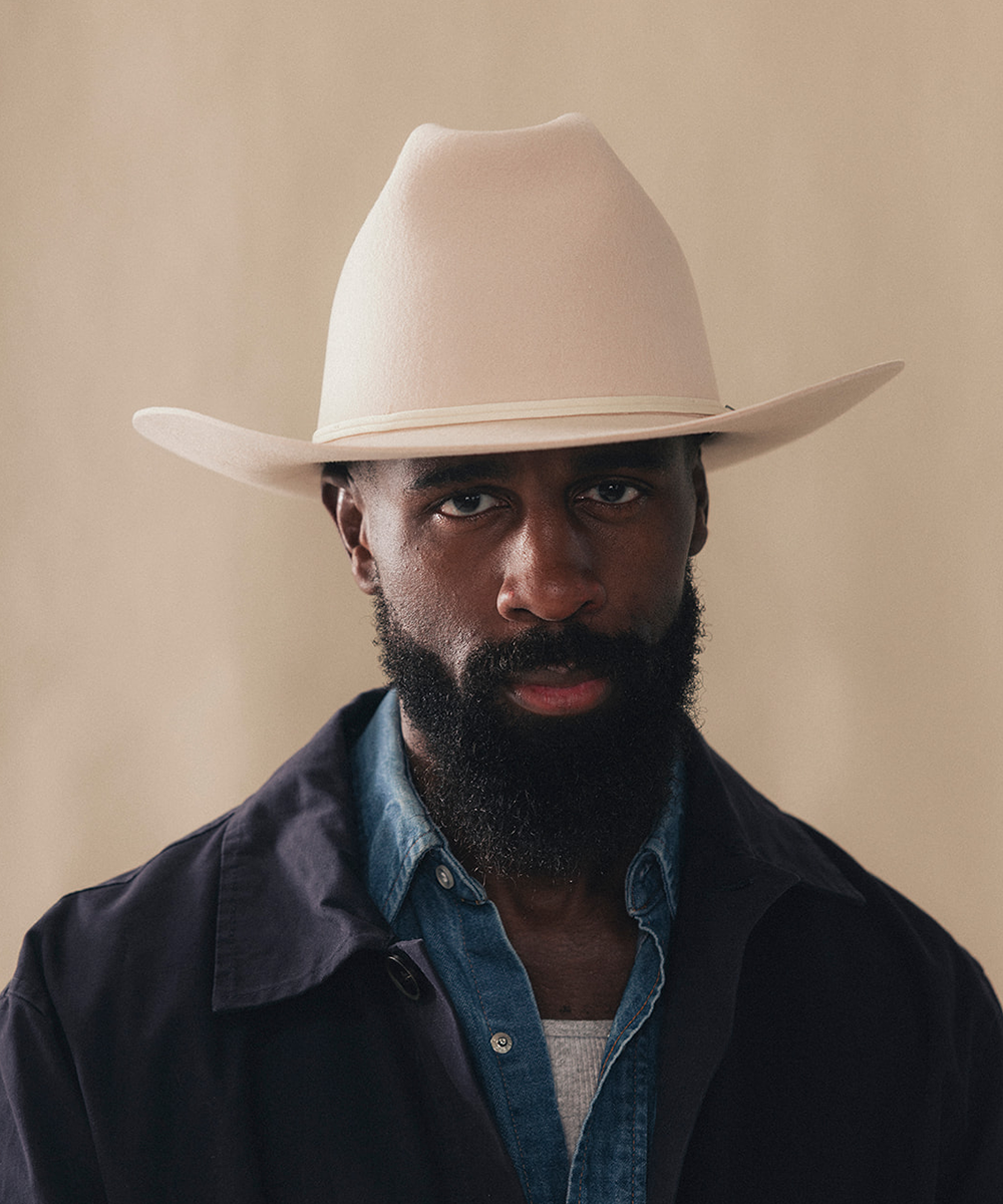 A close-up portrait of a man with a beard wearing a light beige cowboy hat, a blue denim shirt, and a dark jacket, set against a solid beige background #color_buckskin