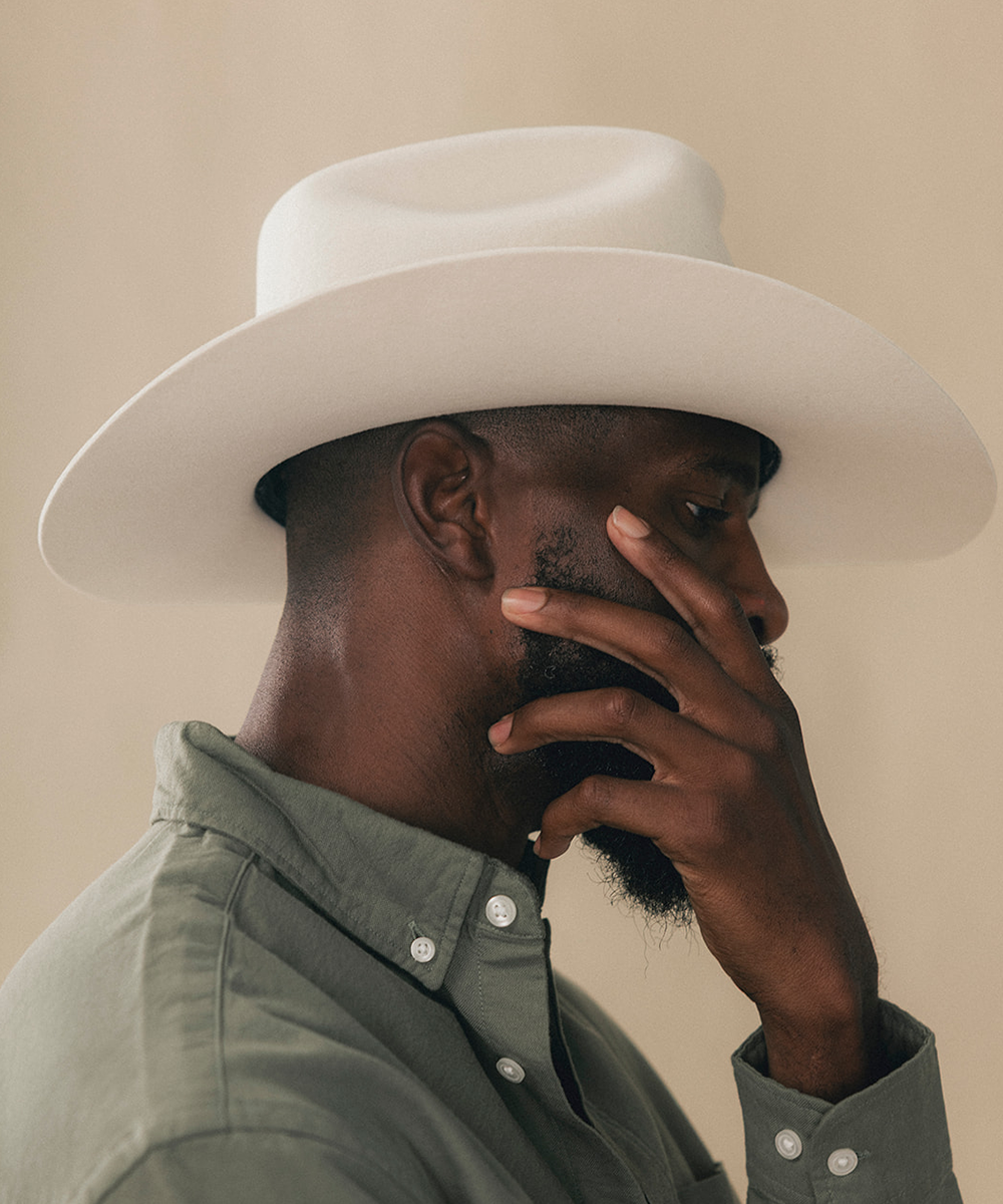 A man in profile wearing a wide-brimmed white cowboy hat and an olive green button-down shirt, with his hand resting against his face, set against a solid beige background #color_bone