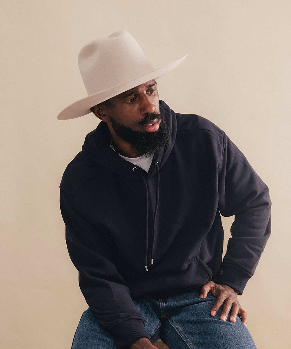 A man wearing a cream-colored wide-brim fedora hat, a dark navy pullover hoodie, and blue jeans, posed against a solid beige studio background #color_silverbelly