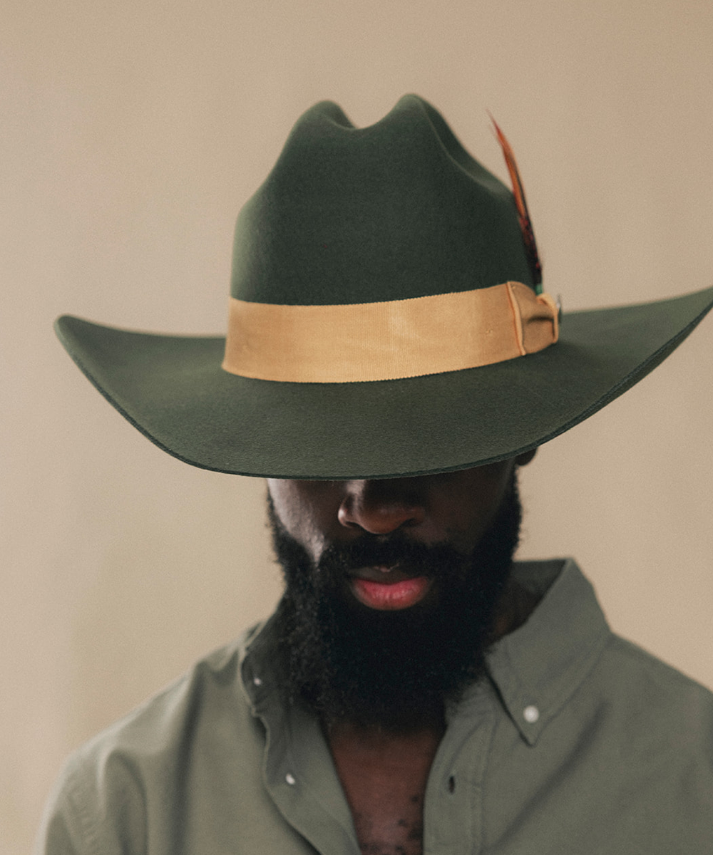 A man wearing a dark green felt cowboy hat with a tan leather band and feather accent, paired with an olive green shirt, against a plain beige background #color_forest green/mustard