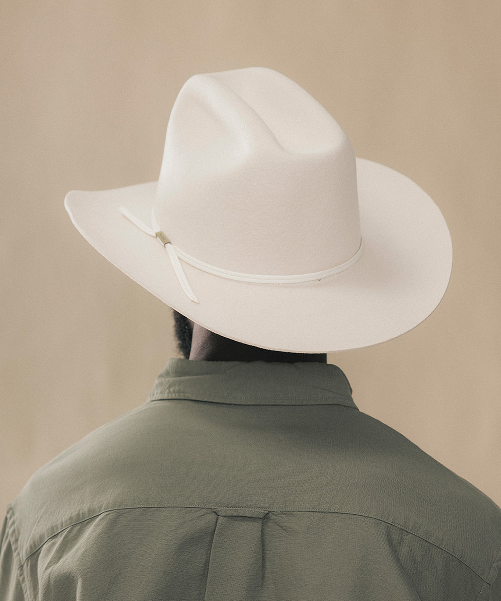 A person shown from behind wearing a white wide-brim cowboy hat with a matching white band and a muted olive green shirt, set against a plain beige background #color_bone