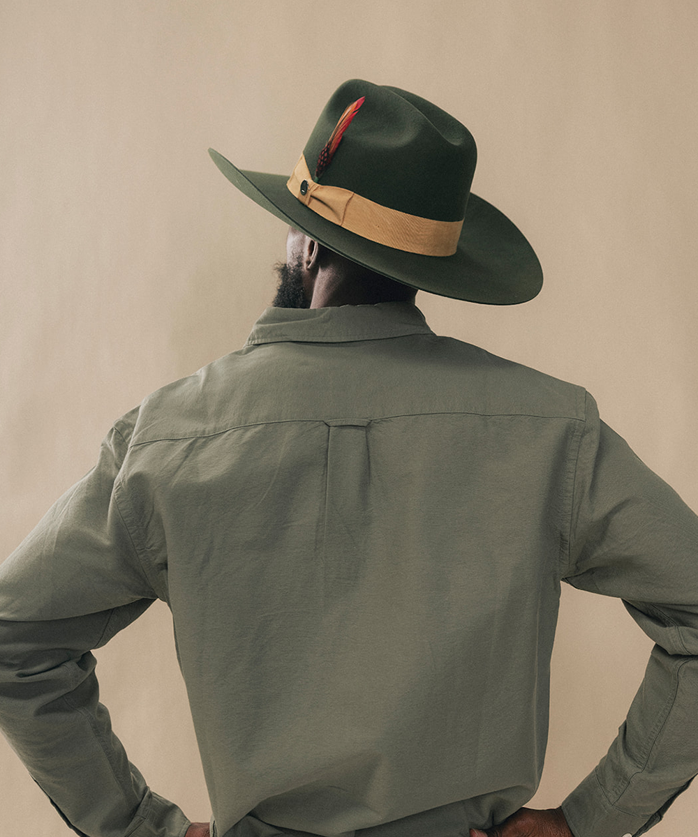 A man viewed from behind wearing a dark green wide-brim fedora hat with a tan band and red feather accent, paired with an olive green shirt against a plain beige studio background #color_forest green/mustard
