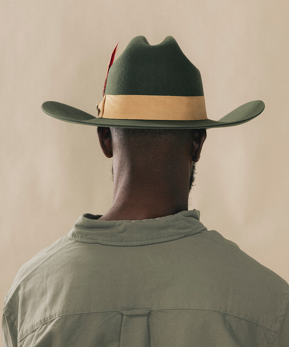 A man viewed from behind wearing a dark green felt cowboy hat with a tan band and a small red feather accent, paired with an olive green shirt against a plain beige background #color_forest green/mustard
