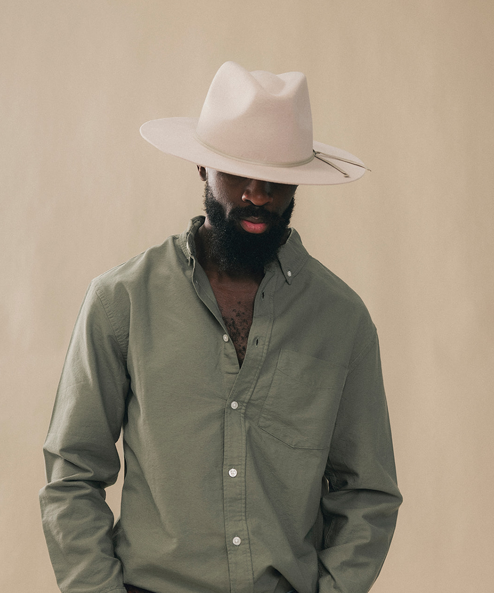 A man wearing a light beige wide-brim fedora hat and an olive green long-sleeve button-down shirt, posed against a solid beige background #color_silverbelly