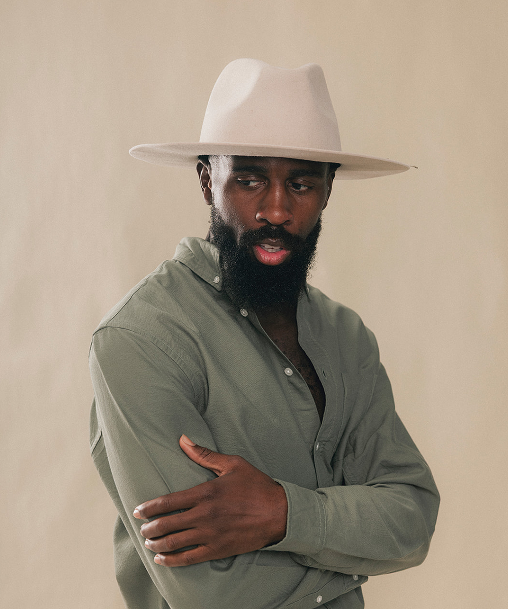 A man with a beard wearing a light beige wide-brim fedora hat and an olive green button-down shirt, posed against a solid beige background #color_silverbelly