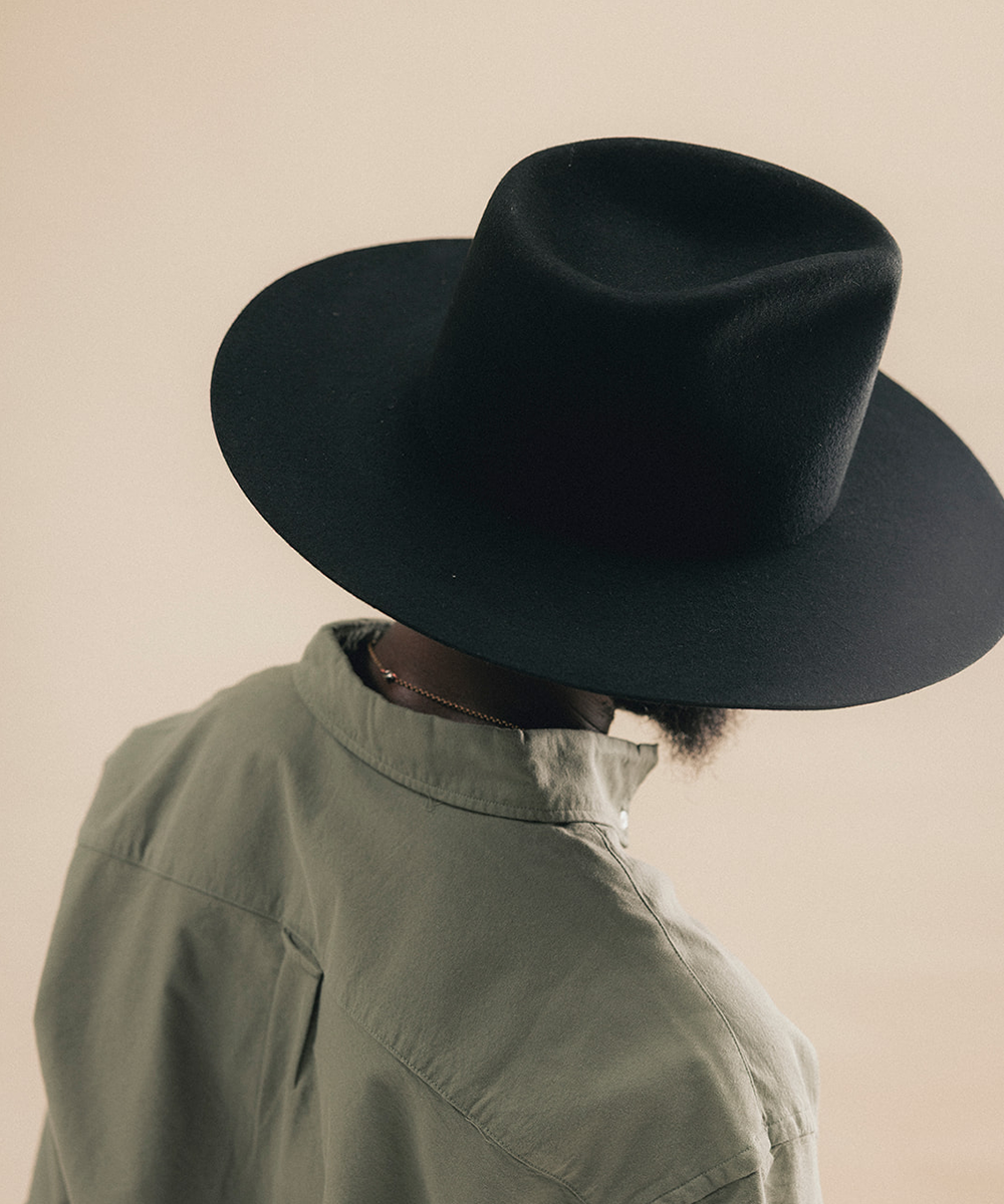 A person shown from the back wearing a wide-brim black hat and an olive green shirt against a plain beige background #color_black