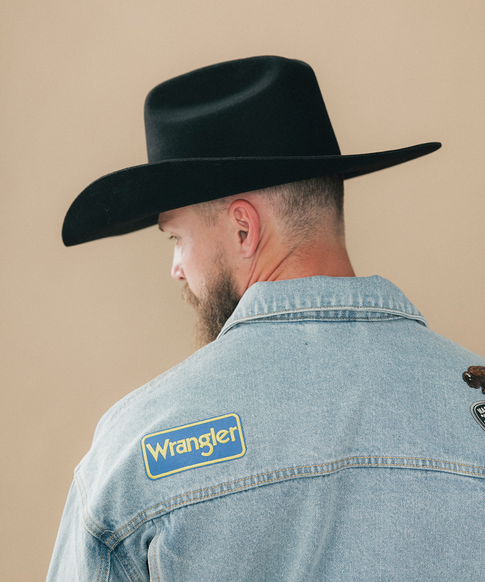 A man wearing a black felt cowboy hat and a denim jacket, viewed from behind against a solid beige background #color_black