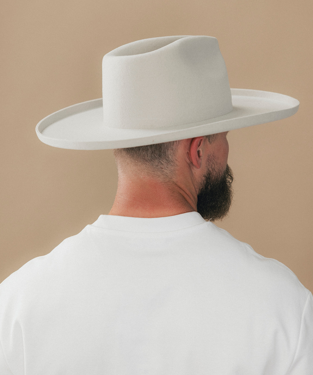 A man wearing a white felt wide-brim fedora hat and a white t-shirt, viewed from behind against a solid beige background #color_silver grey