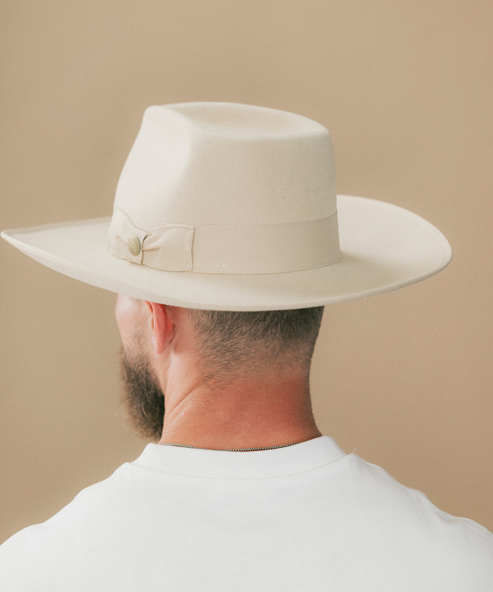 A man viewed from behind wearing a cream-colored fedora hat with a matching band and bow, set against a solid beige background #color_buckskin