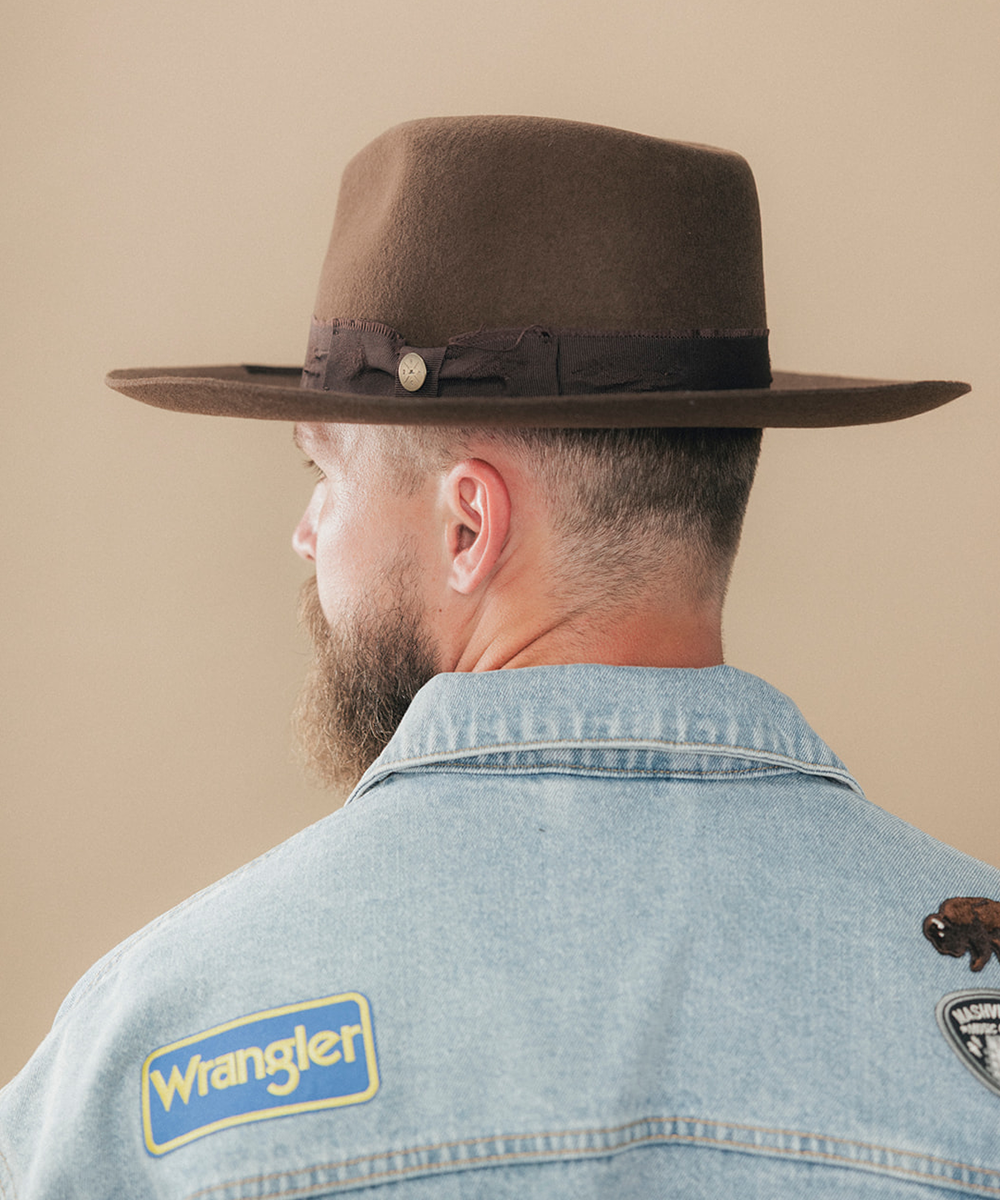 A man wearing a dark brown felt fedora hat and a denim jacket, viewed behind against a solid beige background #color_dark brown