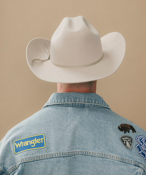 A man wearing a cream felt cowboy hat with a decorative band and a denim jacket, viewed from behind against a solid beige background #color_silverbelly